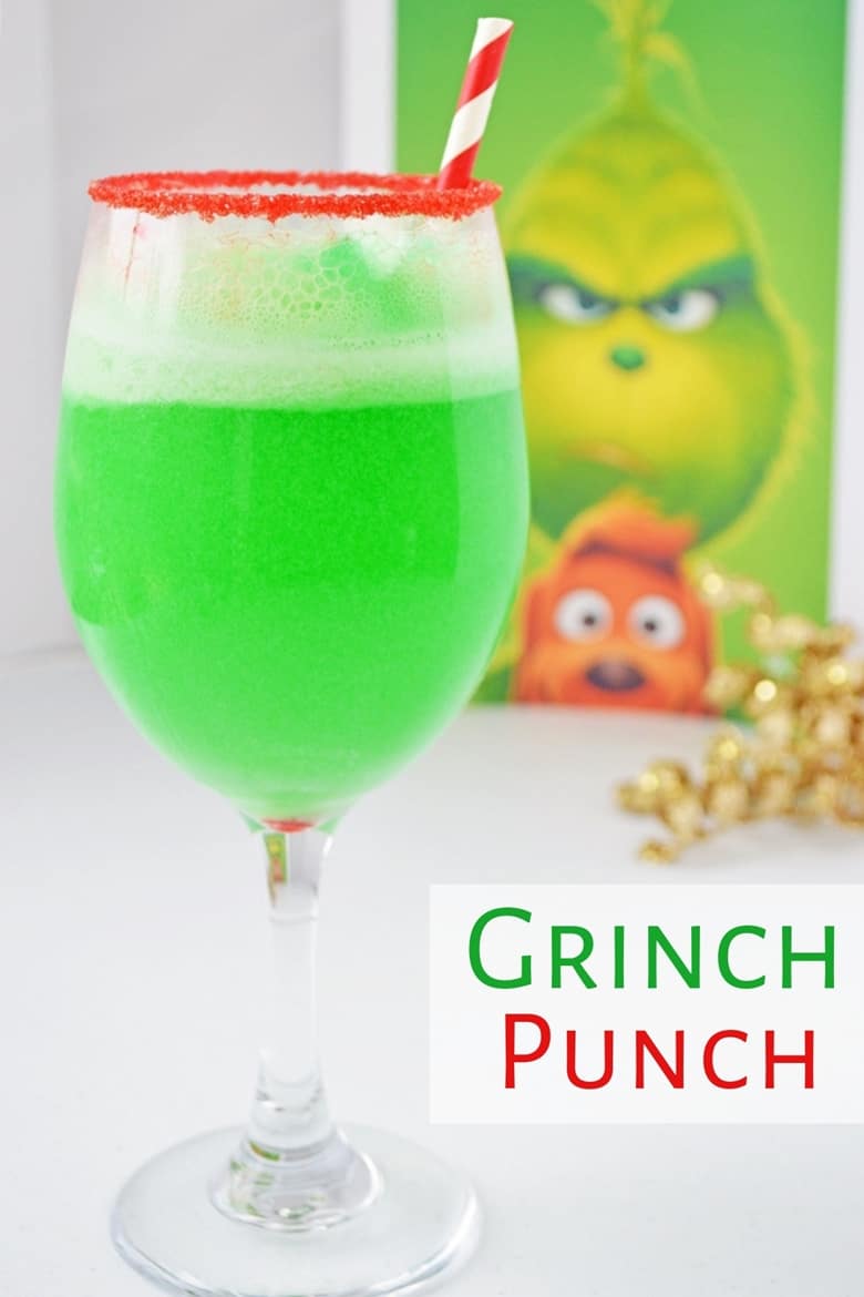 Bright green grinch punch in a red sugar rimmed wine glass with a picture of The Grinch behind it.