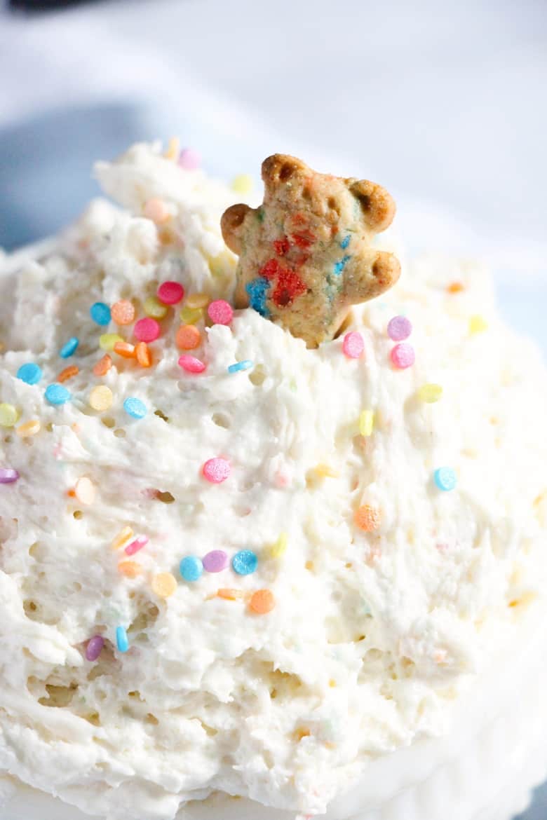 Creamy funfetti dip topped with pastel sprinkles with a teddy bear shaped cookie sticking out of it.