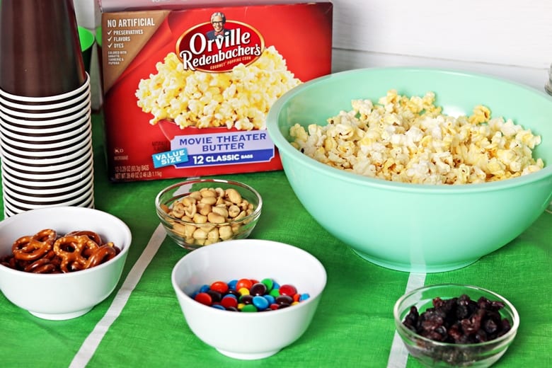 Box of Orville Redenbacher's Popcorn, bowl of popcorn, brown cups, peanuts, chocolate candies, pretzels, and raisins