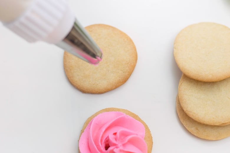 Piping pink icing onto circular sugar cookies, starting at center. A decorated and 3 undecorated cookies are also in the shot.