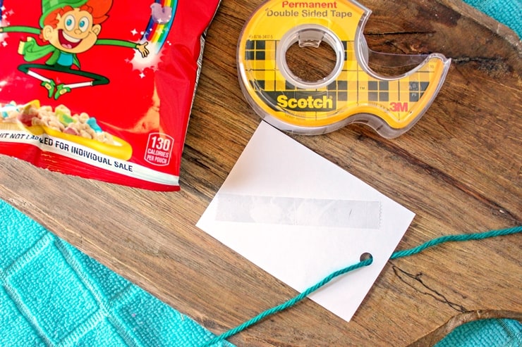 A gift tag turned over with tape on the back and yarn through the hole on top. Roll of double sided tape and a bag of Lucky Charms can be seen next to the tag.