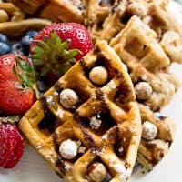 Reese's Puffs Cereal Waffles broken into quarters and served on a white plate with fresh berries, chocolate syrup, and Reese's Puffs.