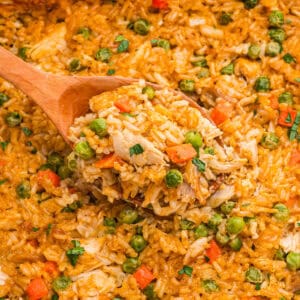 A wooden spoon lifting a scoop of creamy chicken, rice, peas, and carrots from the dish.