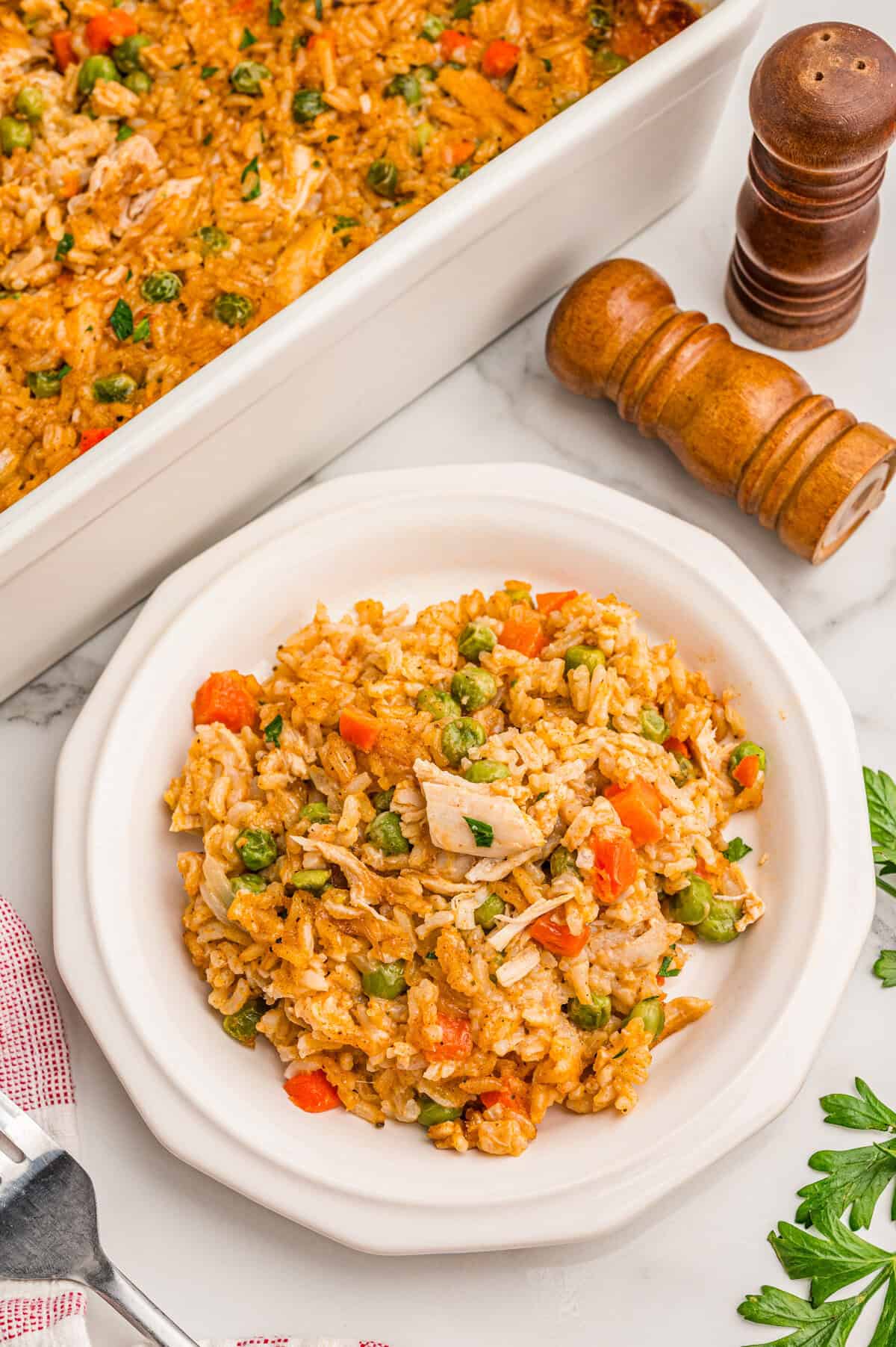 Overhead view of a plated serving of chicken and rice casserole next to the full chicken and rice bake with parsley and salt and pepper shakers.