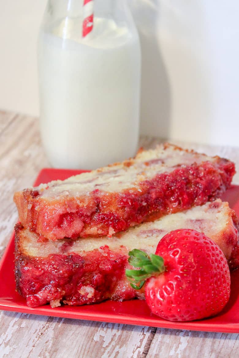 2 slices of strawberry pound cake on a red plate with a strawberry on the side. A glass of milk with a red and white straw is in background.