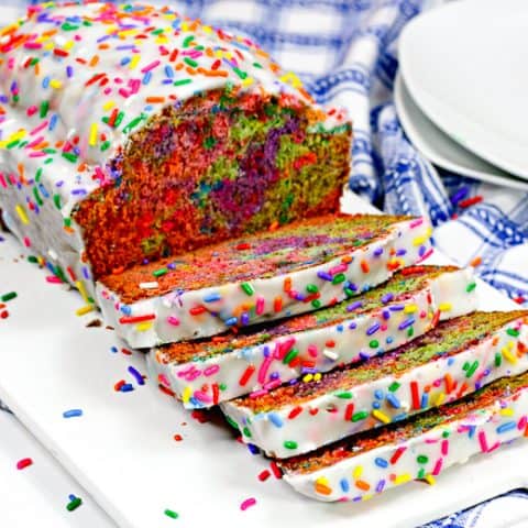 Rainbow sprinkle covered iced unicorn bread on white cutting board, with 4 slices cut