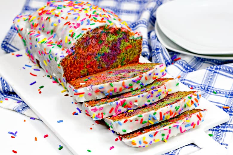 Rainbow sprinkle covered iced unicorn bread on white cutting board, with 4 slices cut.