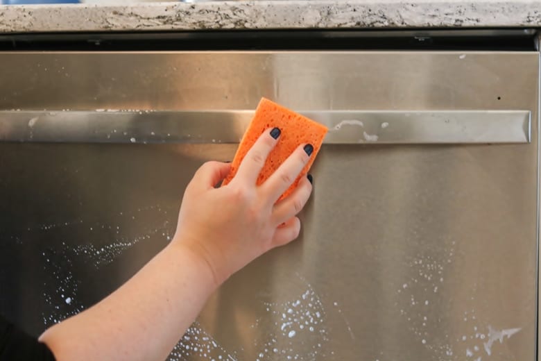 A hand holding a sponge and wiping a sudsy stainless steel dishwasher handle