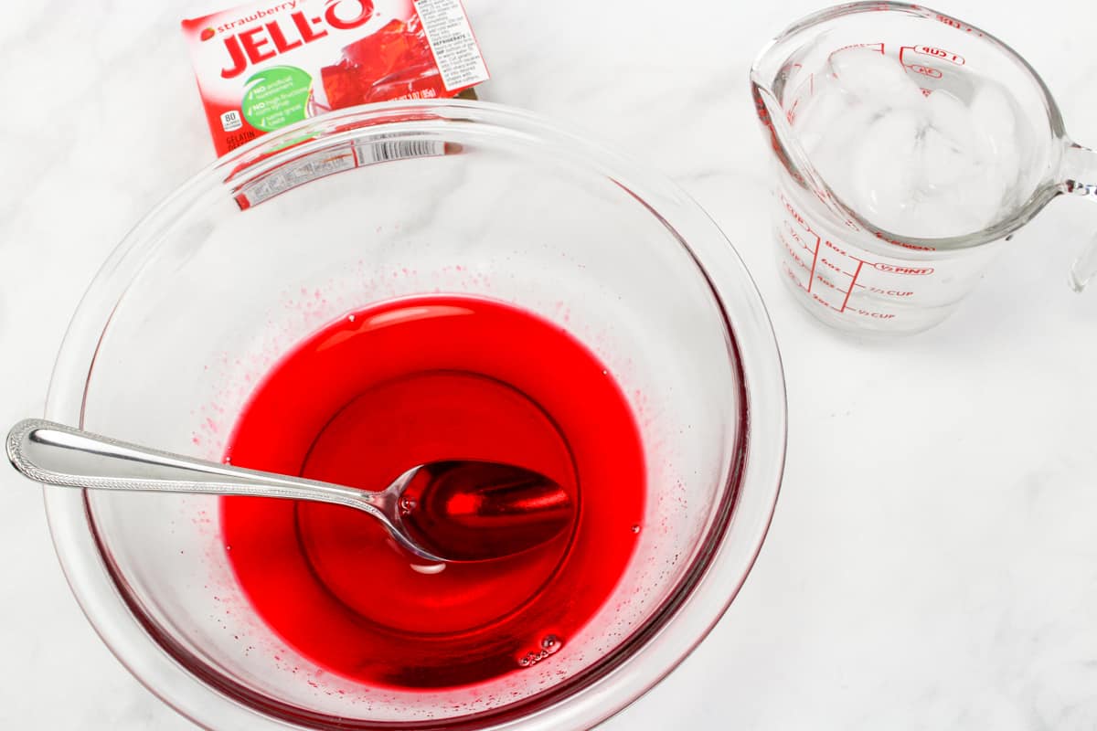 Bowl of jello mix dissolved in water with a spoon in it and box of strawberry jell in background.