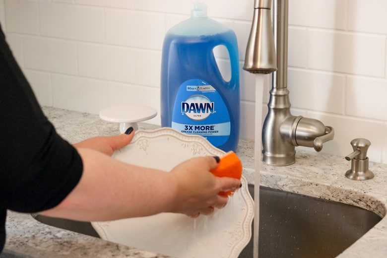 Someone washing a white plate in a kitchen sink. Bottle of Dawn dish soap is in background.