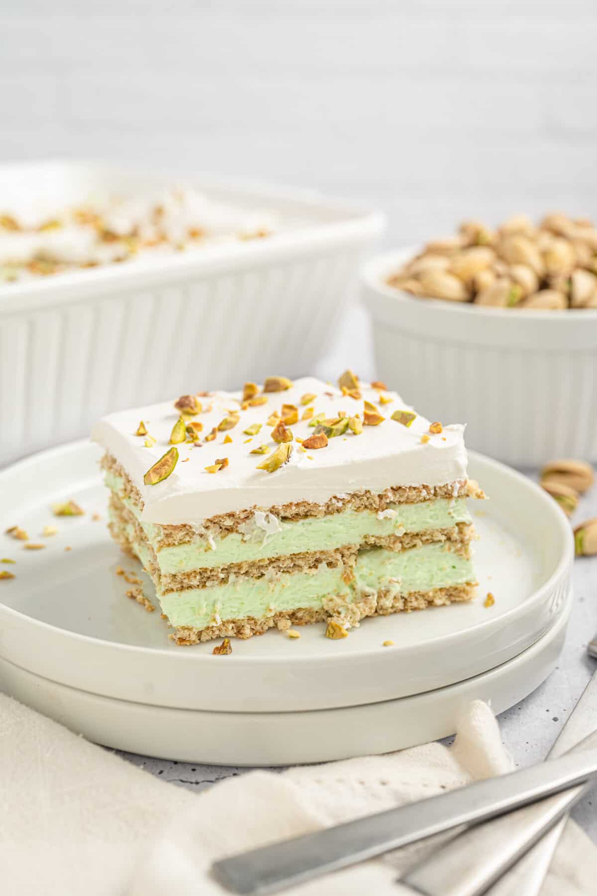A slice of pistachio icebox cake sits on a white plate, layered with graham crackers, green pistachio filling, and whipped topping with pistachio garnish. In the background, a bowl of whole pistachios and the larger baking dish of cake are visible, creating a cozy dessert scene.