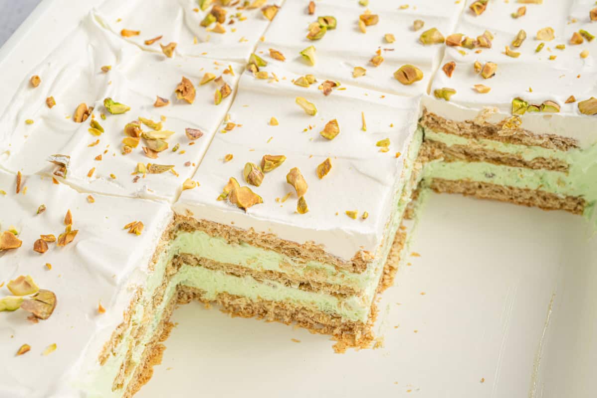 Whole pistachio icebox cake with a few slices removed, exposing the neat layers of graham crackers and green pistachio filling beneath a smooth whipped topping sprinkled with chopped pistachios.