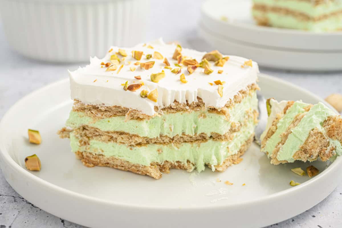 A slice of pistachio icebox cake sits on a white plate, featuring visible layers of graham crackers and pistachio-flavored filling, topped with whipped cream and crushed pistachios. A fork with a bite-sized piece rests nearby, and another plate with cake is slightly blurred in the background.