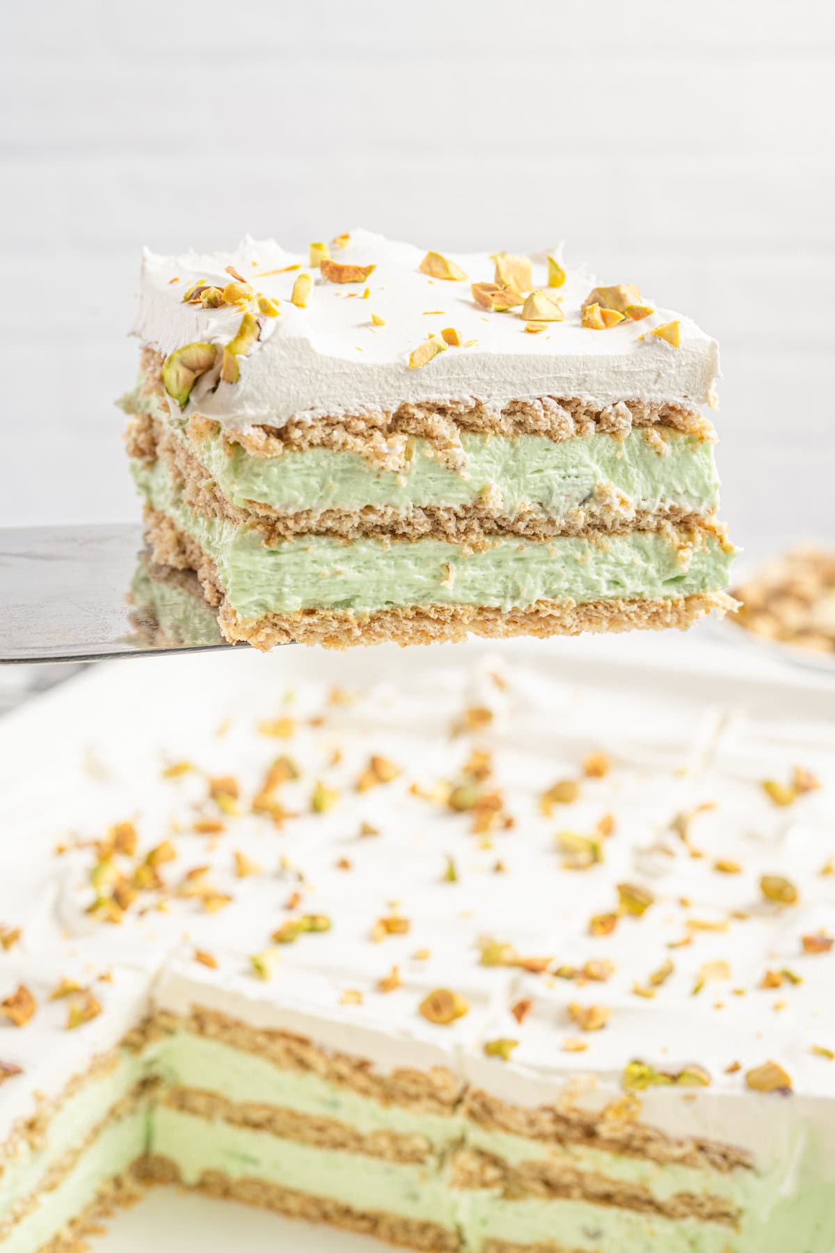 A spatula lifts a single slice of no bake pistachio icebox cake from a larger dish, showing clean layers of graham crackers, green pistachio pudding filling, and cool whip with crushed pistachios. The remaining cake is visible below.