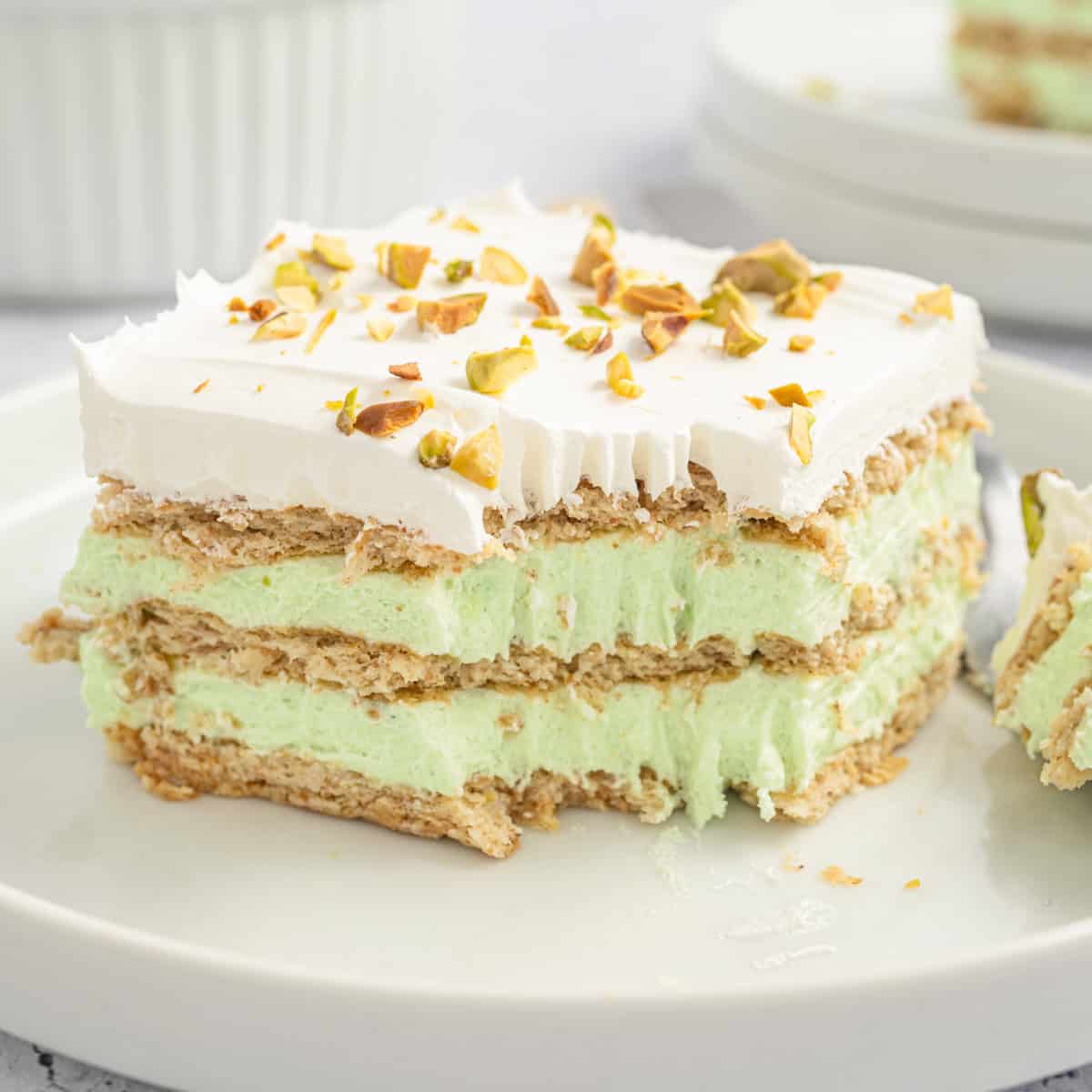 A slice of pistachio icebox cake with layers of graham crackers, green pistachio filling, and whipped topping, garnished with chopped pistachios.
