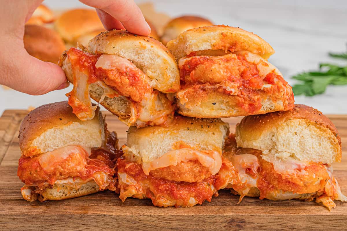 Chicken parmesan sliders are stacked on a wooden board. Each sandwich contains a breaded chicken nugget, marinara sauce, and melted cheese, with slightly toasted, herb-sprinkled buns. A hand is lifting one from the stack.