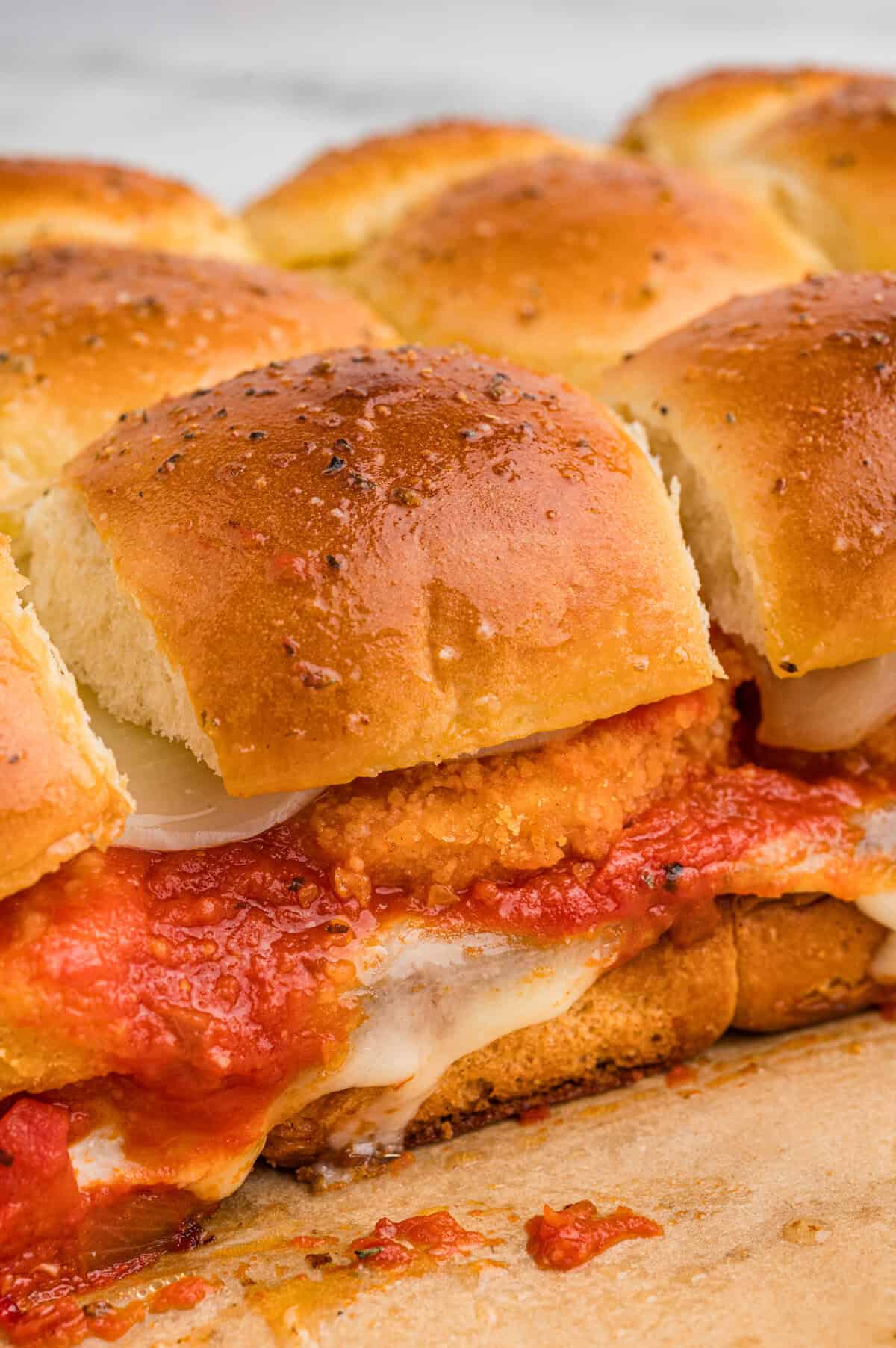 Close-up of golden brown chicken parm sliders with nuggets in a row, showing layers of breaded chicken, marinara sauce, and melted mozzarella between soft, herb-topped buns.