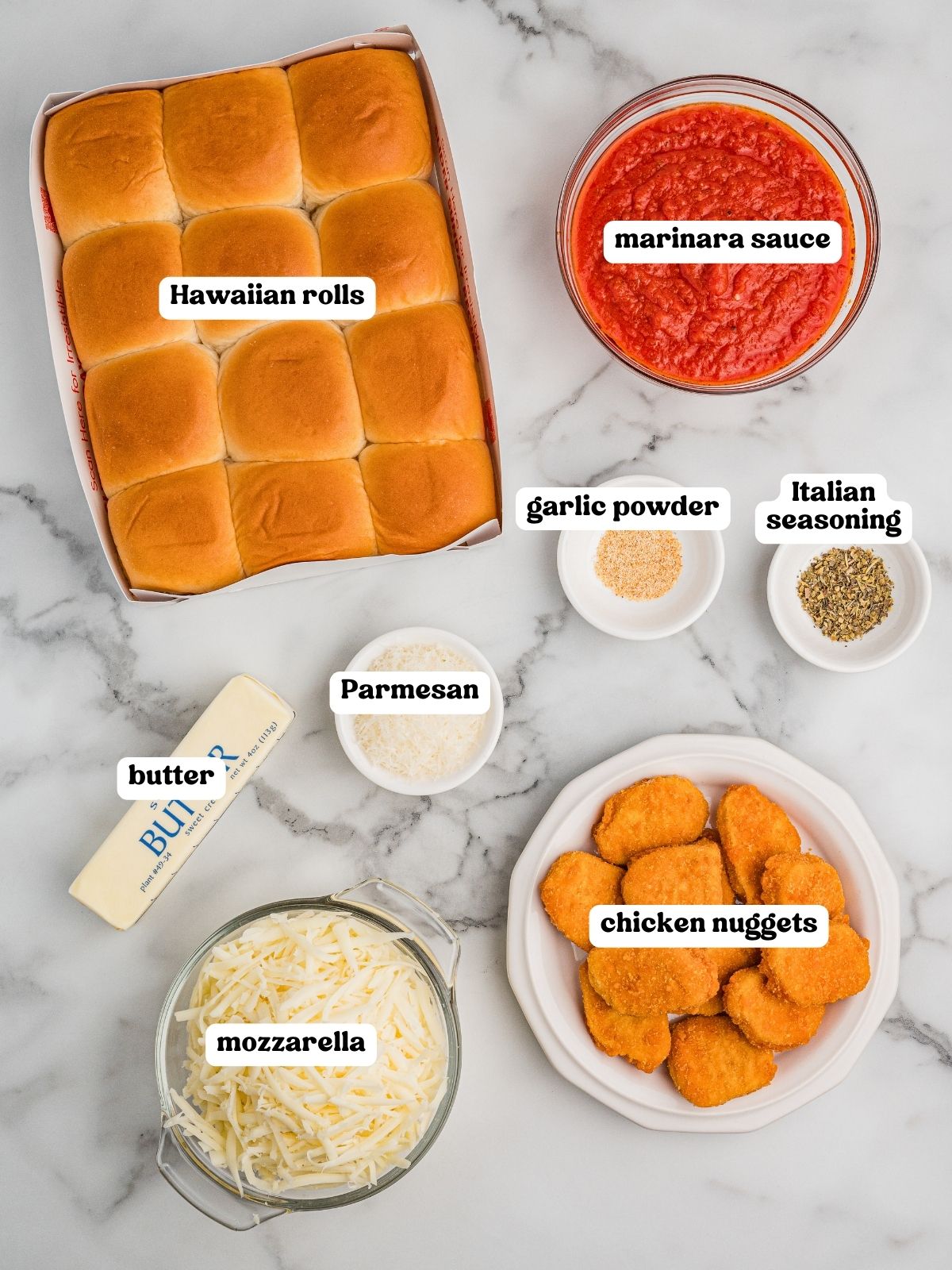 Labeled ingredients for chicken parm sliders on a marble surface for chicken parmesan sliders, including a package of Hawaiian rolls, a bowl of marinara sauce, a stick of butter, a bowl of shredded mozzarella, a bowl of grated Parmesan, a plate of chicken nuggets, and small bowls of garlic powder and Italian seasoning.
