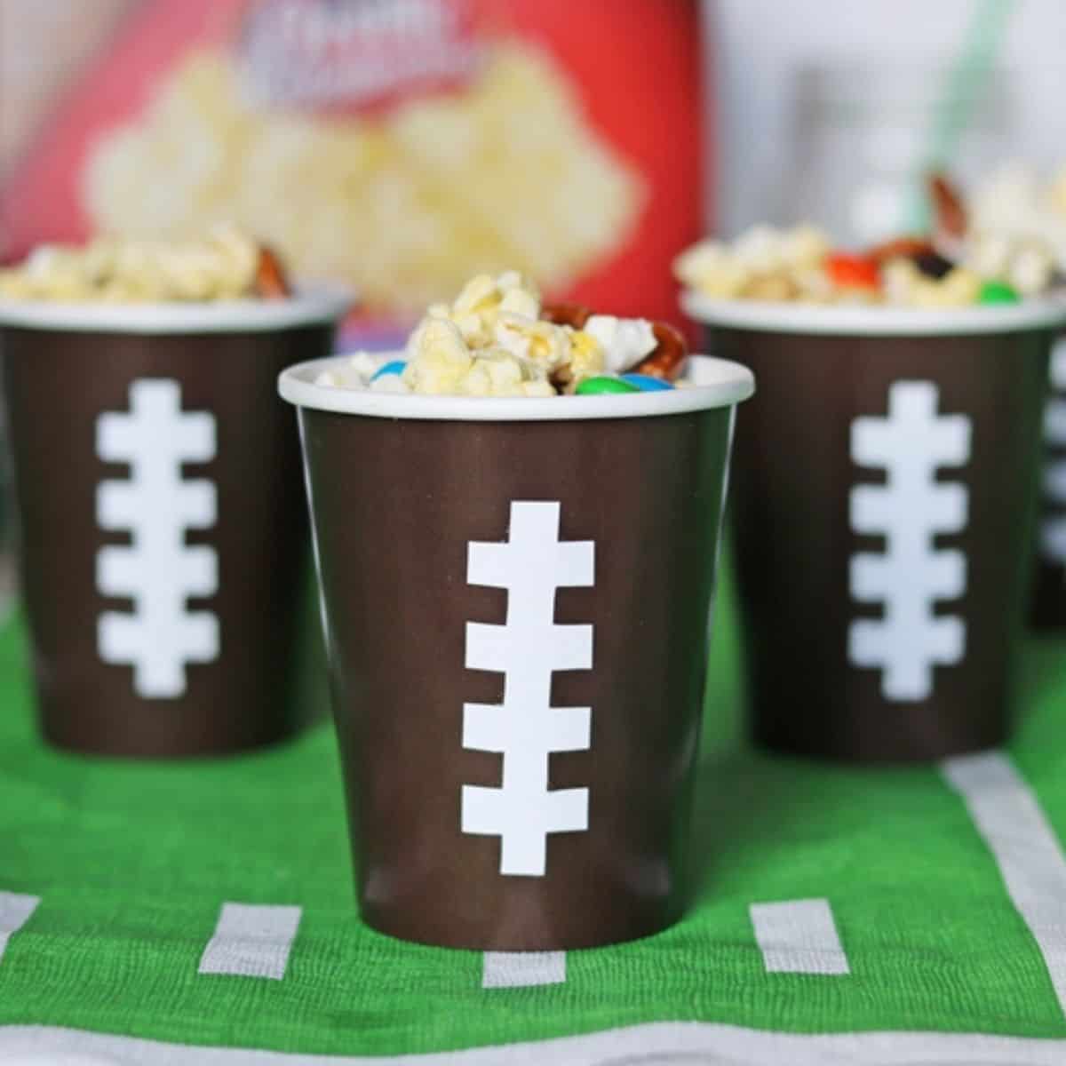 Snack mix in football cups with popcorn, pretzsels, and M&Ms.
