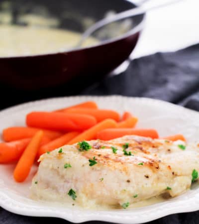 Chicken breast and baby carrots on white dinner plate, with skillet filled with creamy sauce in the background