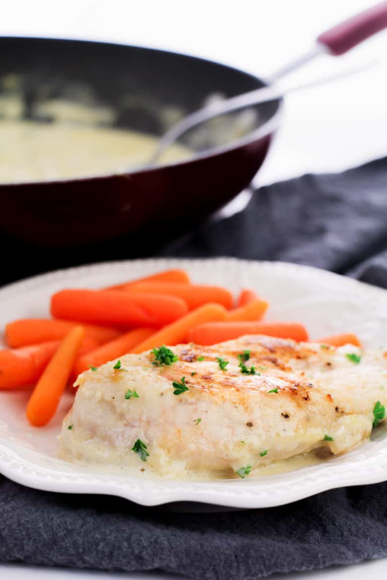 Chicken breast and baby carrots on white dinner plate, with skillet filled with creamy sauce in the background