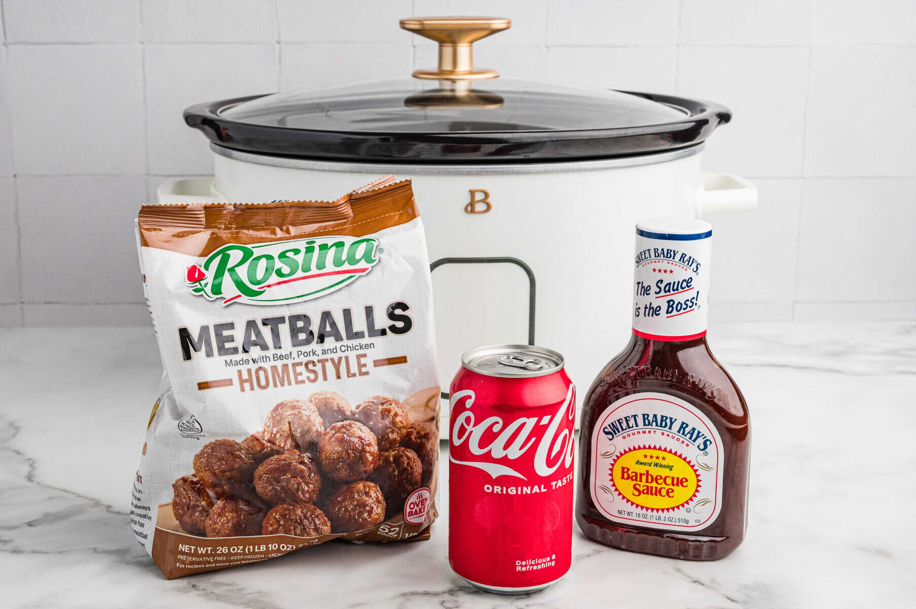 A package of Rosina Homestyle Meatballs, a can of Coca-Cola, and a bottle of Sweet Baby Ray&rsquo;s Barbecue Sauce are arranged in front of a white slow cooker on a marble countertop.