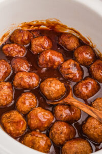 Coca-Cola meatballs in slow cooker with a wooden spoon.