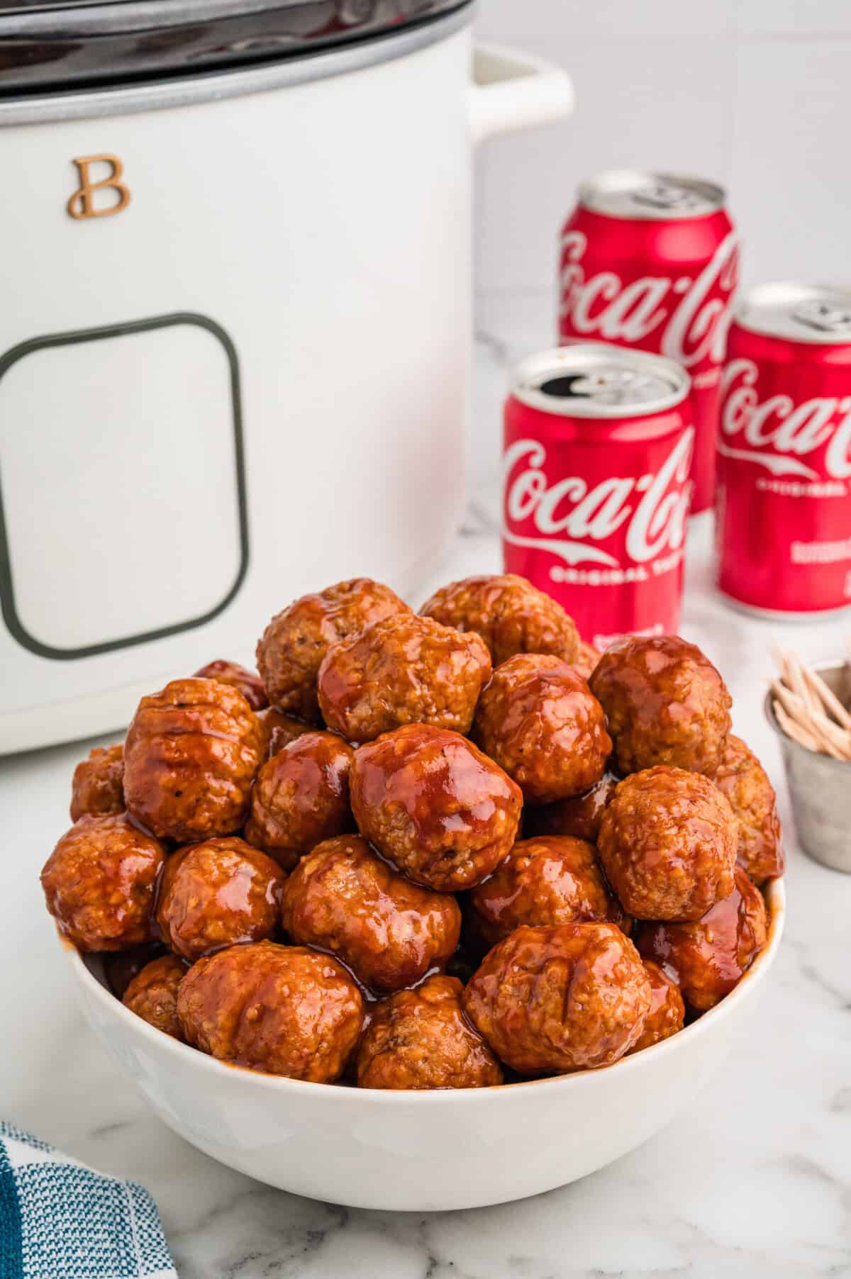A bowl of crockpot coca cola meatballs sits in front of a white slow cooker and coke cans.