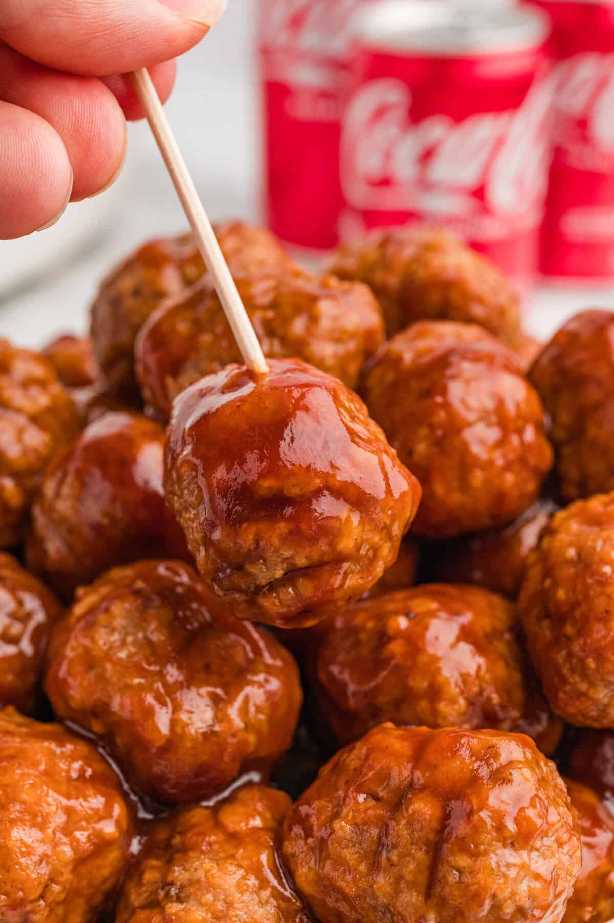 A hand holding a coca cola meatball on a toothpick above a pile of similarly glazed meatballs. Blurred red Coca-Cola cans are visible in the background.