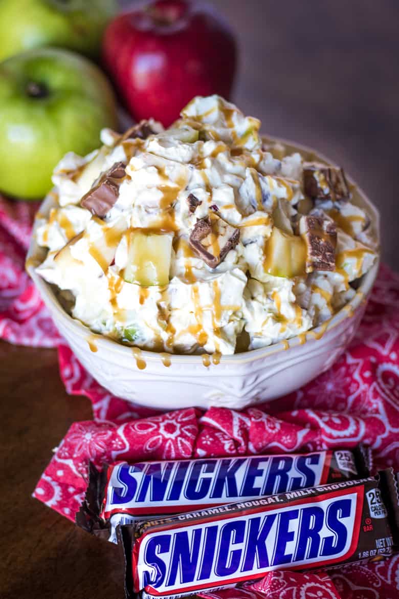 Snickers Apple Salad in a white bowl, 3 apples in background and 2 Snickers candy bars in foreground.