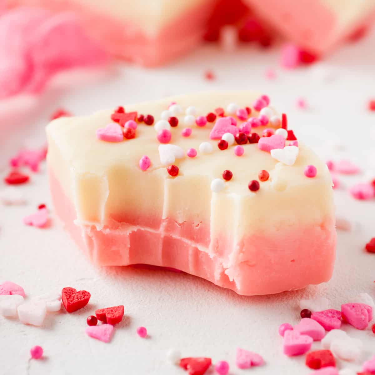Piece of Valentines fudge with pink and white layers and topped with festive pink and red sprinkles. A bite has been taken out of the fudge.