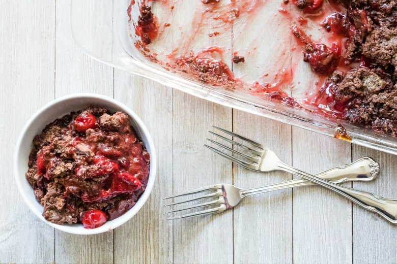 Small serving of chocolate cherry dump cake in an individual bowl with forks and remaining cake in glass pan beside it.