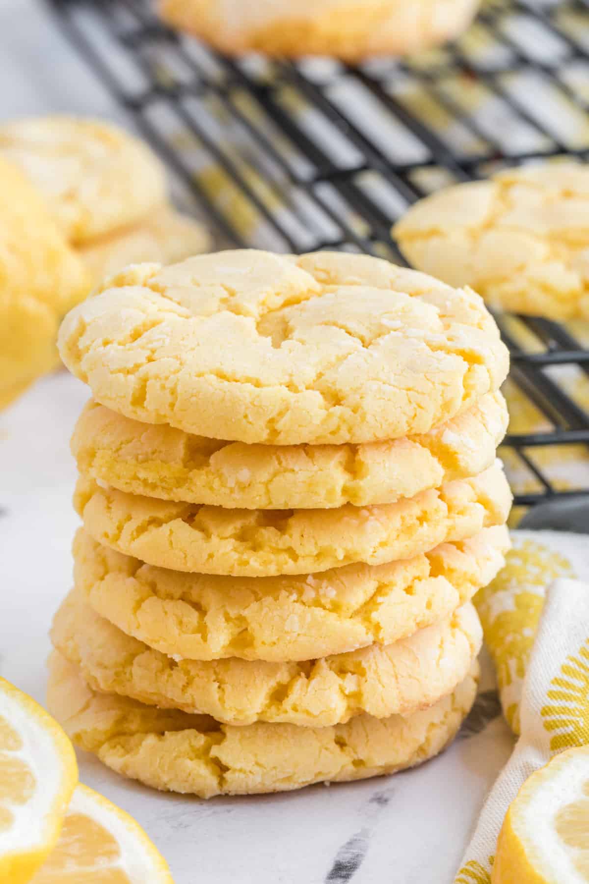 Lemon cake mix cookies stacked on top of each other with cookies cooling on a rack behind them.