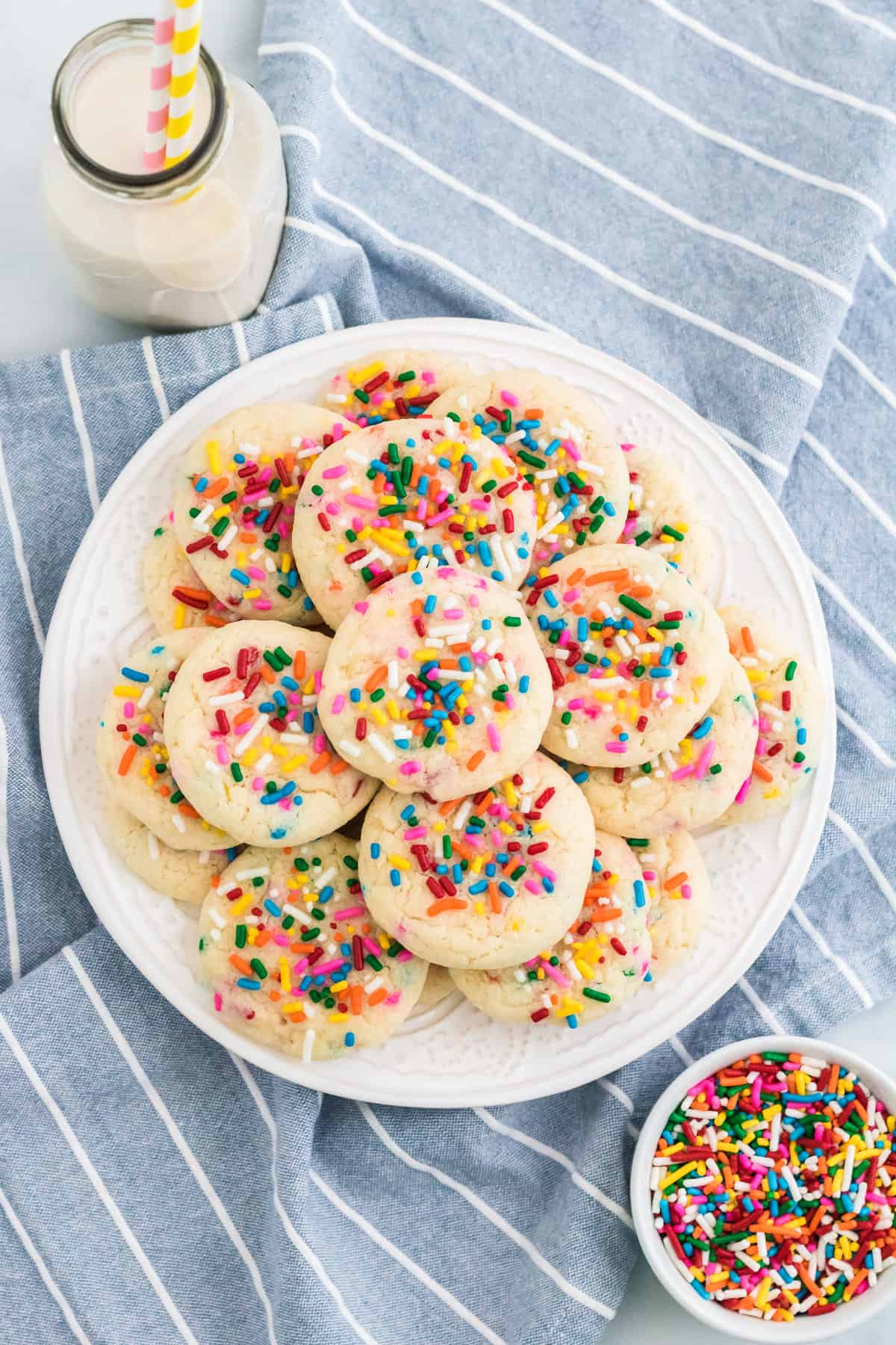 Funfetti cake mix cookies with rainbow sprinkles on a white plate with glass of milk and extra sprinkles beside them.