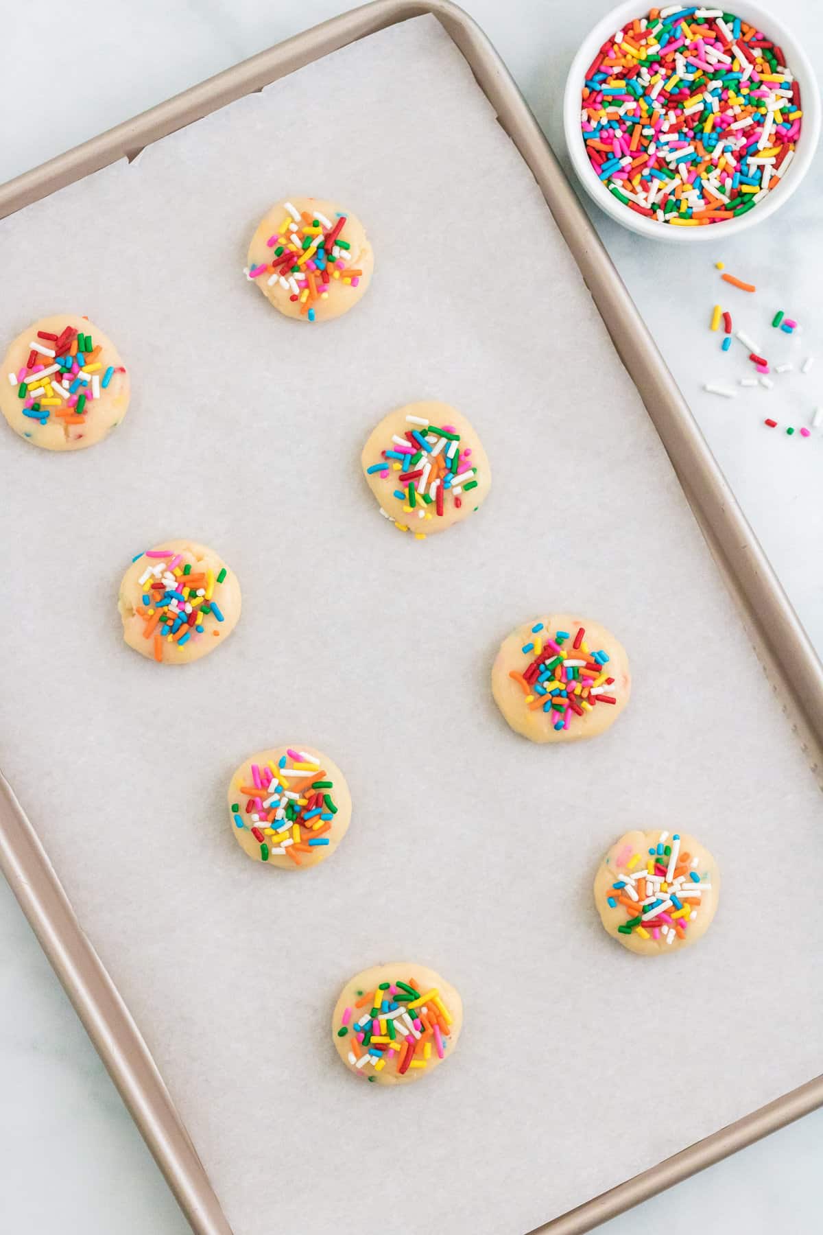 Rainbow sprinkled topped Funfetti cookie dough on lined baking sheet. The dough is pressed down slightly into hockey-puck shaped discs. 
