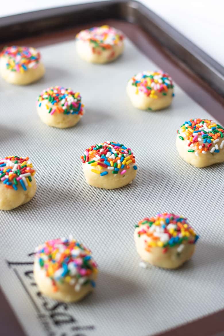 Cookie dough balls topped with rainbow sprinkles on silicone mat lined backing sheet.