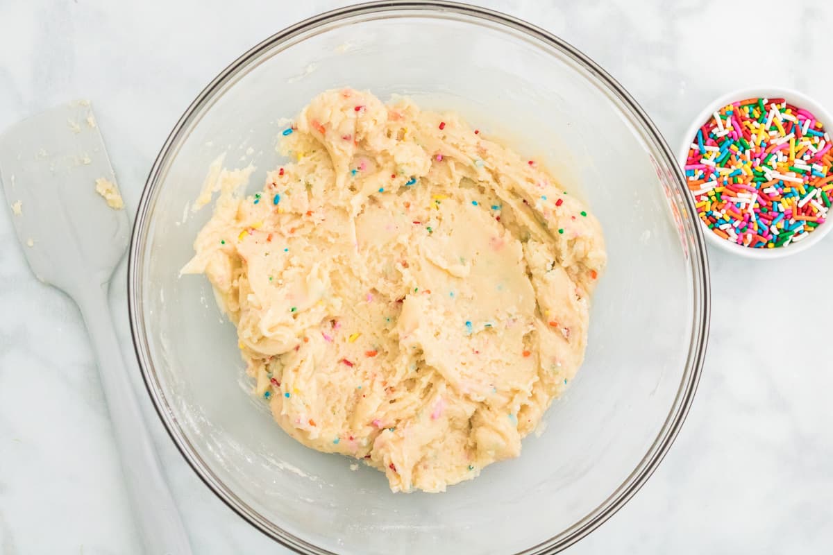 Funfetti cake mix cookie dough in a large mixing bowl with rainbow sprinkles next to it.