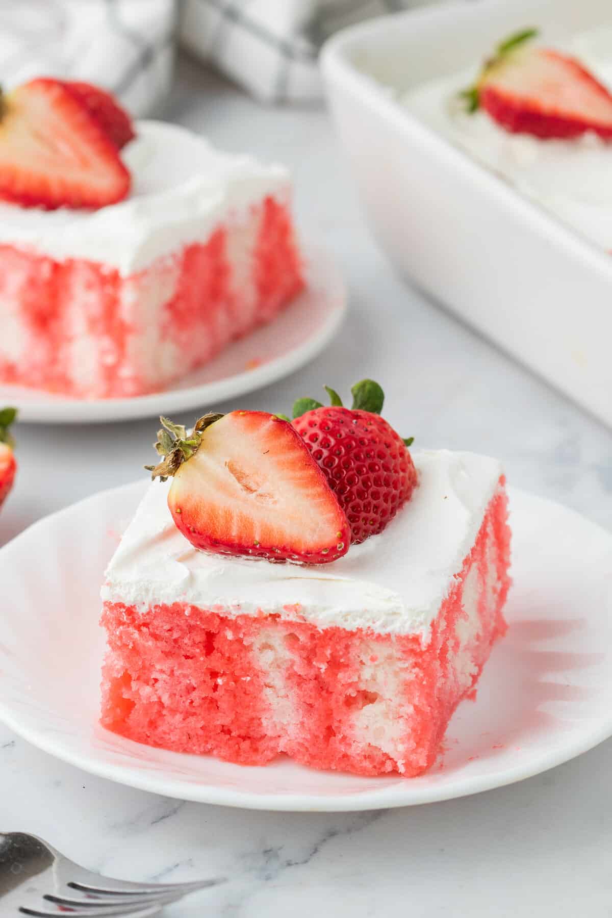 Strawberry jello poke cake topped with cool whip whipped topping and fresh strawberries and served on white plates.
