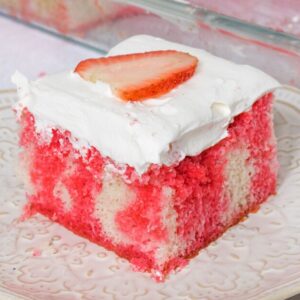 Square slice of strawberry jello poke cake made from white cake with red Jello drips through it. The cake is topped with cool whip topping and a slice of strawberry.