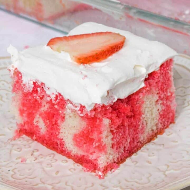 Square slice of white cake with red Jello drips through it. The cake is topped with cool whip topping and a slice of strawberry.
