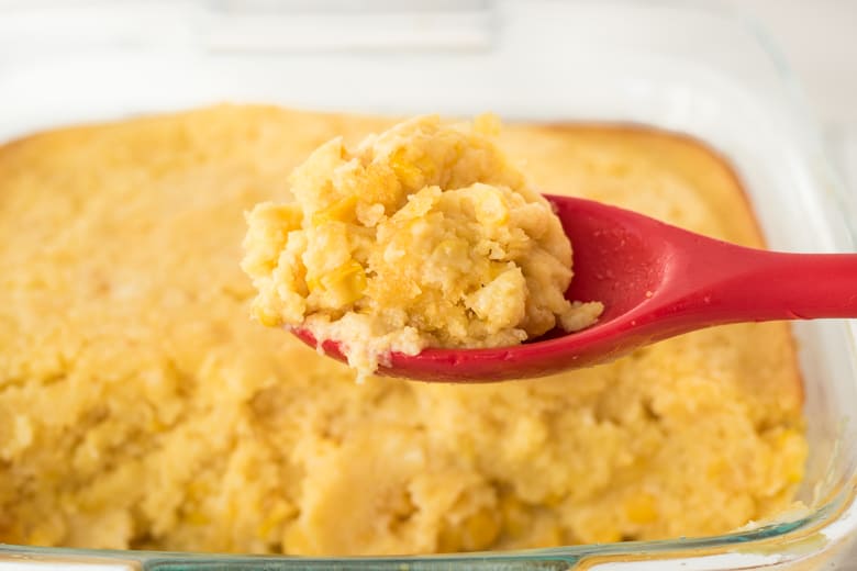 Jiffy corn casserole being scooped from baking pan with a red serving spoon.