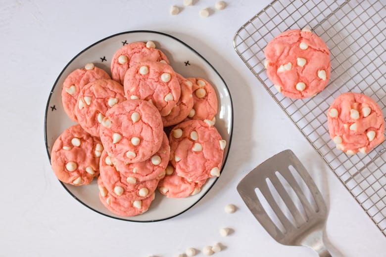 Strawberry cake cookies with white chocolate chips on a plate with a metal spatula and more cookies cooling on wire rack next to it.