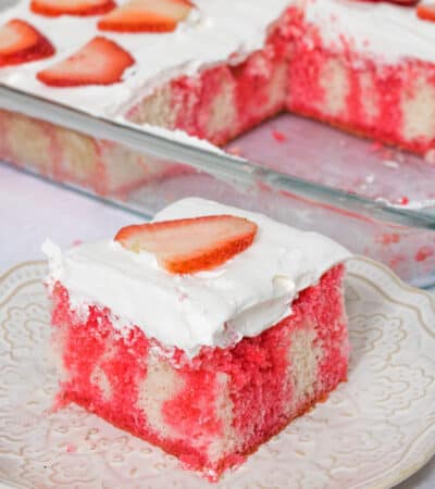 Square slice of white cake with red Jello dripping down through it. The cake is topped with cool whip topping and a slice of strawberry.