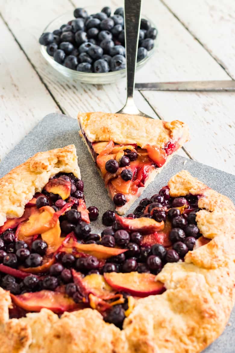 Rustic Galette with peach and blueberry filling. One triangular slice is cut and being served, with a bowl of fresh blueberries in the background.