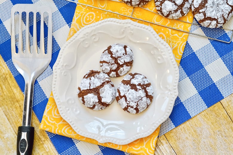 Chocolate crinkle cookies on white plate with spatula and cooling rack covered in cookies in background