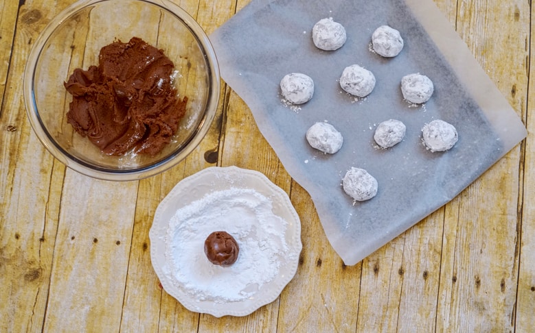 chocolate fudge cookie dough balls being rolled in powdered sugar then placed on lined baking sheet