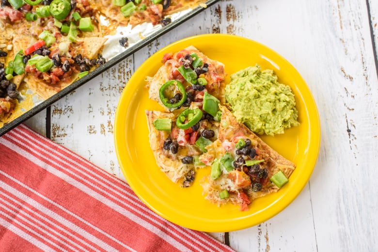 Nachos with beans, peppers, and melted cheese on yellow plate with guacamole on the side. Sheet pan with more nachos in background.