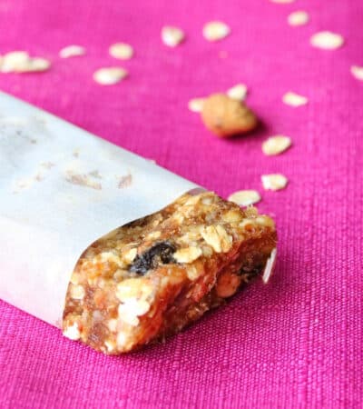 Homemade granola bar wrapped in parchment paper with end exposed. Rolled oats are sprinkled on the pink tablecloth in the background.