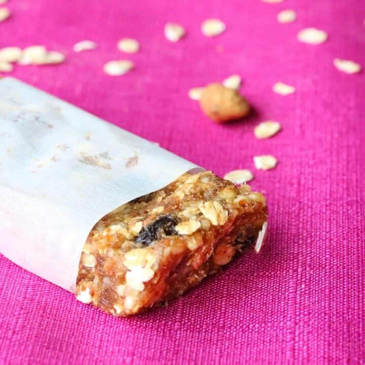 Homemade granola bar wrapped in parchment paper with end exposed. Rolled oats are sprinkled on the pink tablecloth in the background.