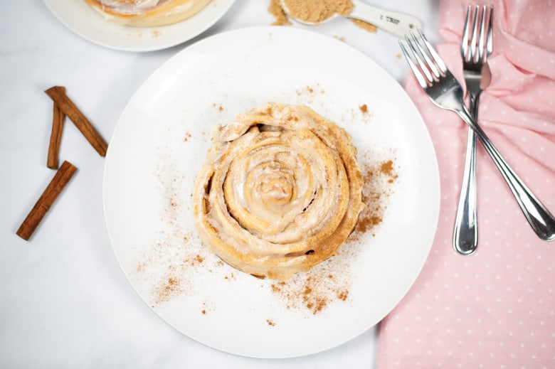 Large cinnamon roll with cream cheese frosting on a white plate with forks and cinnamon sticks next to it